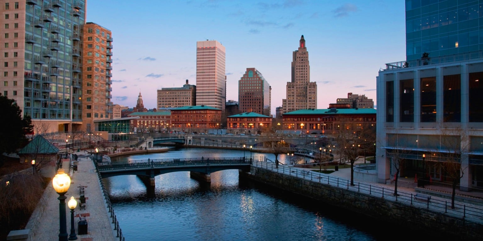 City skyline at dusk with tall buildings, a river, arched bridges, and lamp-posts lining the waterfront. The sky is light blue, and the city lights are starting to come on.
