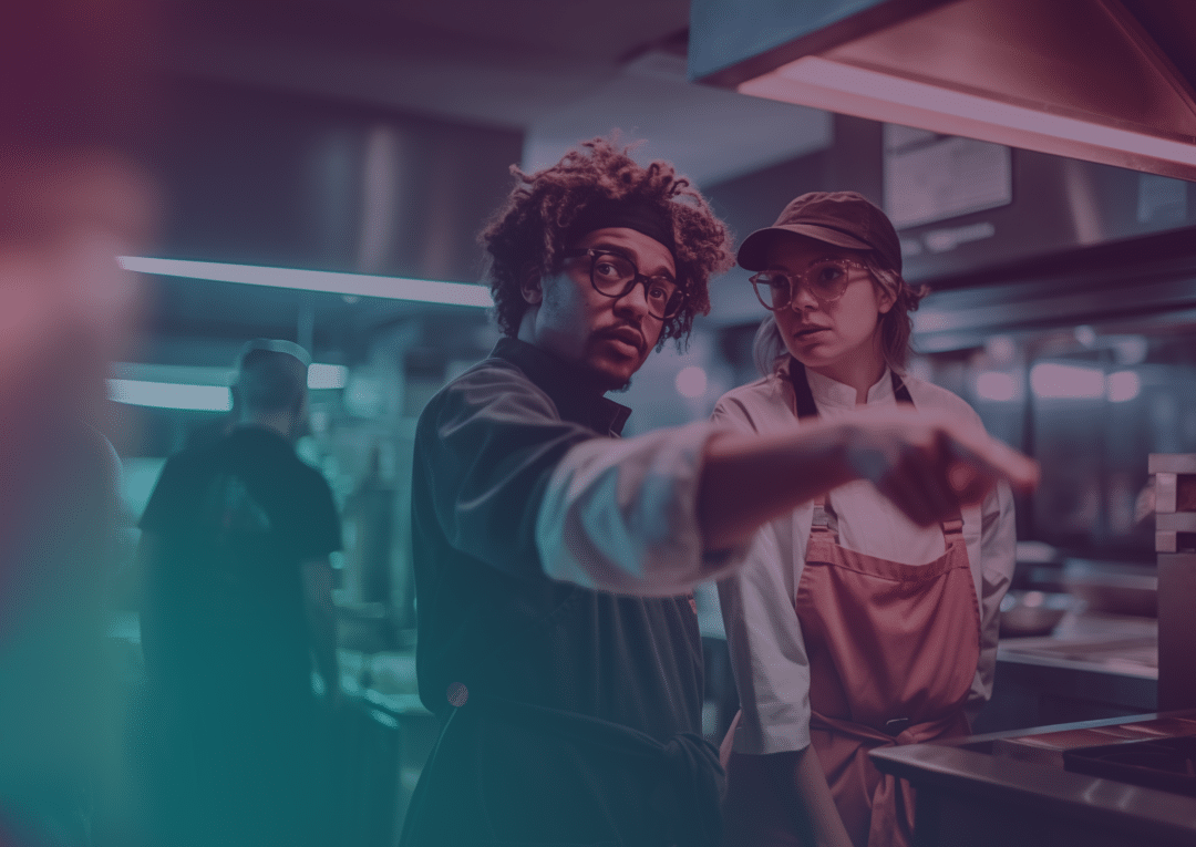 Two chefs in a busy kitchen, one with glasses and curly hair points ahead whilst the other, wearing a hat and apron, listens attentively. The kitchen is lit with blue and pink tones.