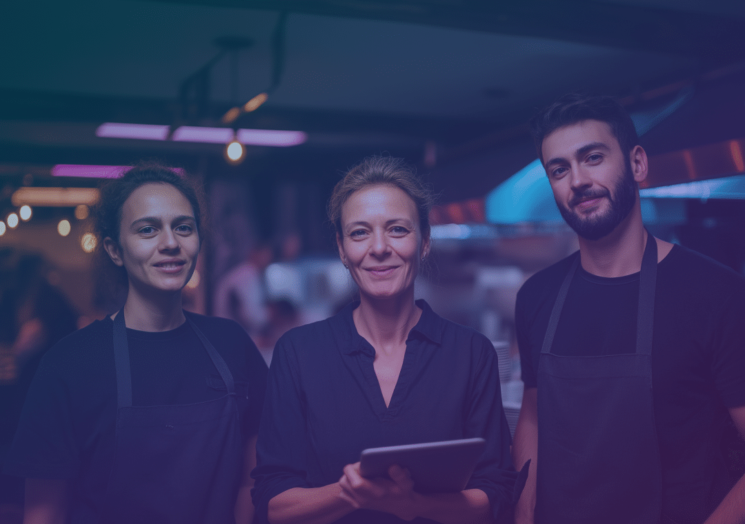 Three restaurant staff members, two women and one man, wearing black aprons, stand together in a modern kitchen. The central woman holds a tablet and all are smiling. The background has warm lighting and blurred kitchen details.