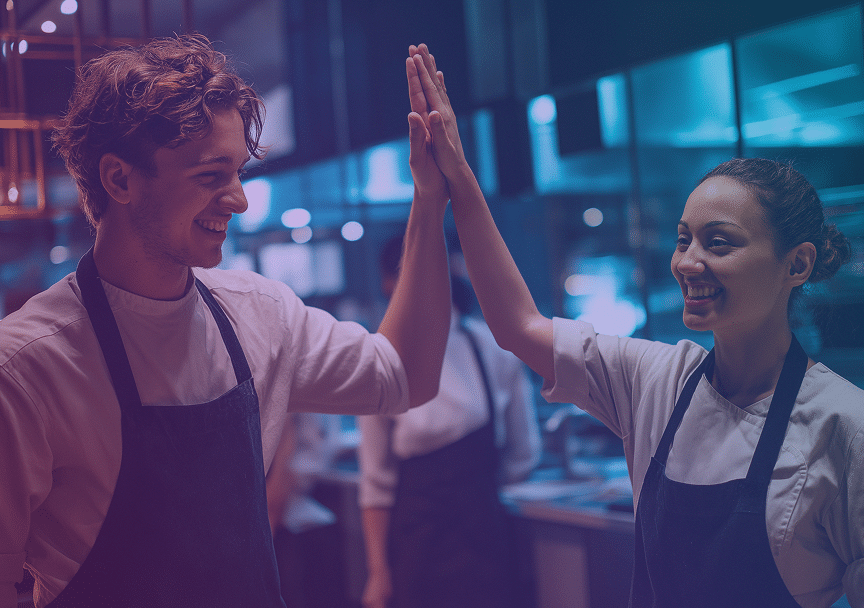 Two restaurant workers in uniforms smile and give each other a high five in a kitchen, celebrating a success.