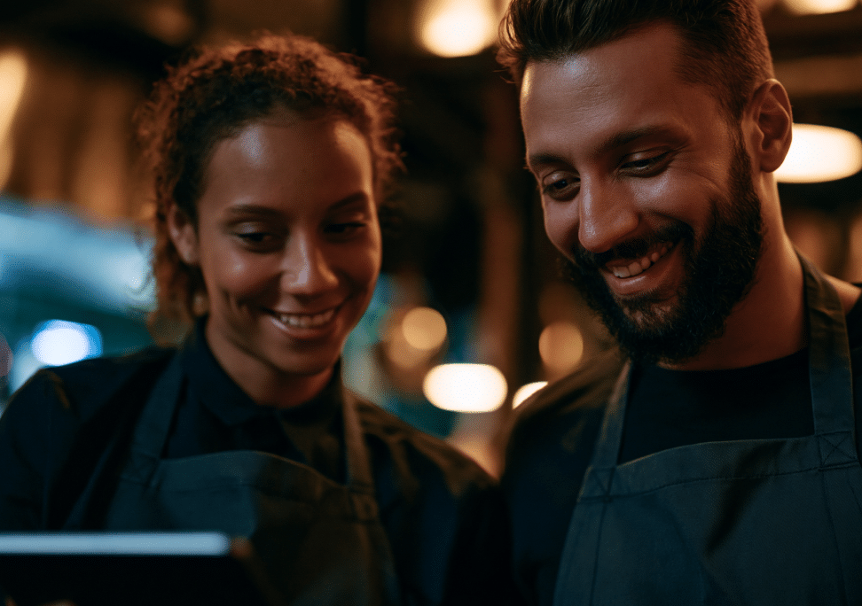 Two people wearing dark aprons smile as they look at a tablet together in a warmly lit restaurant or café setting.