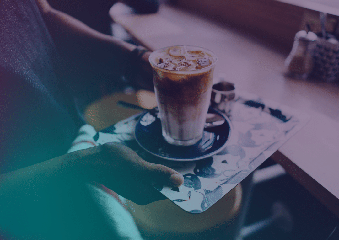 A person holds a tray with an iced coffee in a glass, served on a blue saucer, at a café table by the window.