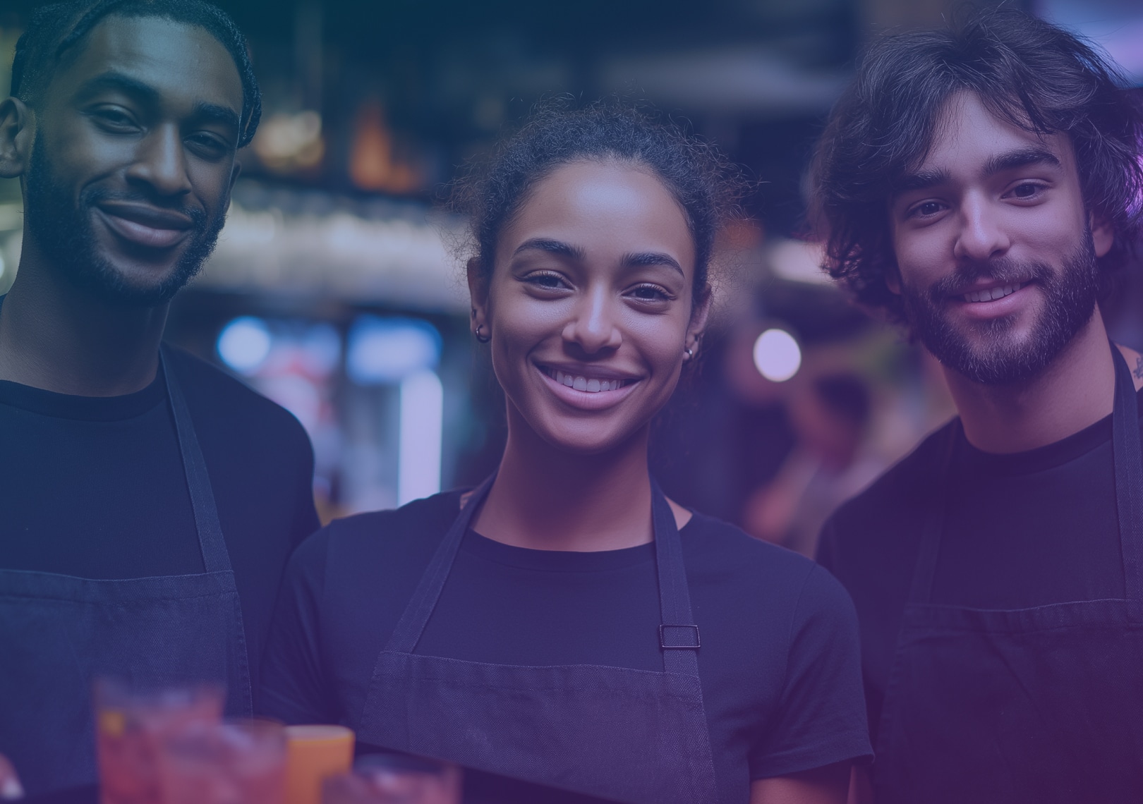 Three people wearing black shirts and aprons smile at the camera in a lively indoor setting, possibly a restaurant or café, with blurred lights and glasses in the background.