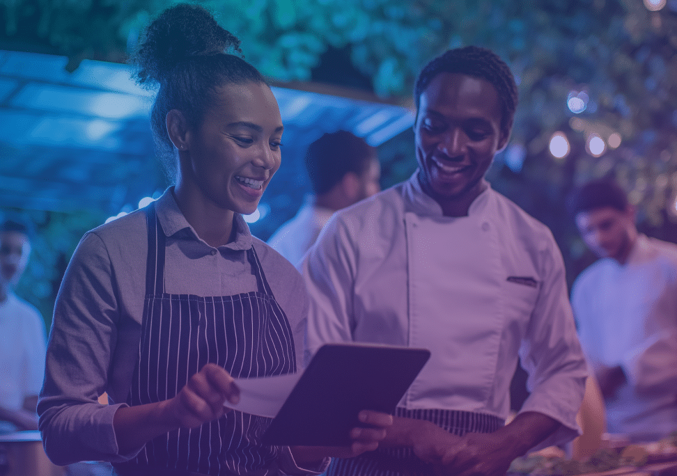 Two people wearing aprons, one holding a tablet and smiling, stand together in an outdoor setting with other people working in the background under fairy lights.