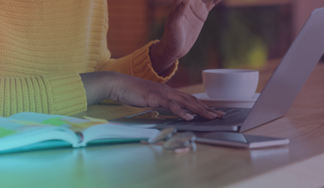 A person in a yellow jumper types on a laptop at a wooden table with an open notebook, a cup, a pair of glasses, and a mobile nearby.