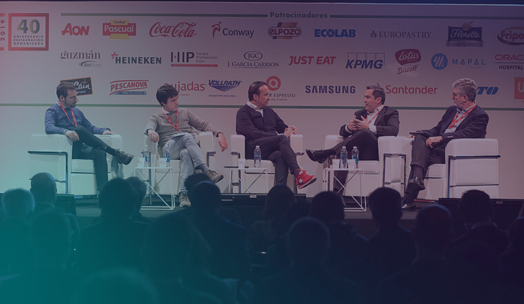Five men sit on stage in white chairs, engaged in a panel discussion at a conference, with sponsor logos displayed on the backdrop. The audience is visible in the foreground.