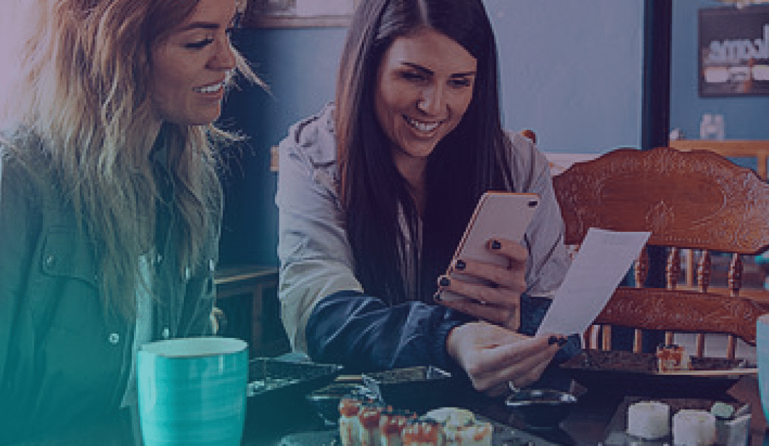 Two women sit at a table with food, smiling as one holds a receipt and a mobile, possibly calculating or splitting the bill. A turquoise mug and plates of food are visible in front of them.