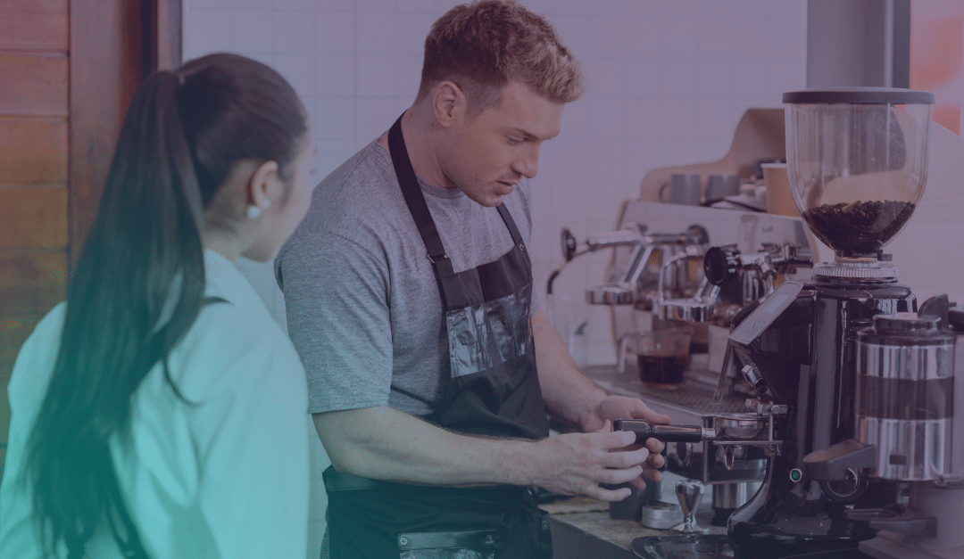 A barista in a grey shirt and apron prepares coffee at an espresso machine while a woman with long dark hair, wearing a white jacket, watches him in a café setting.