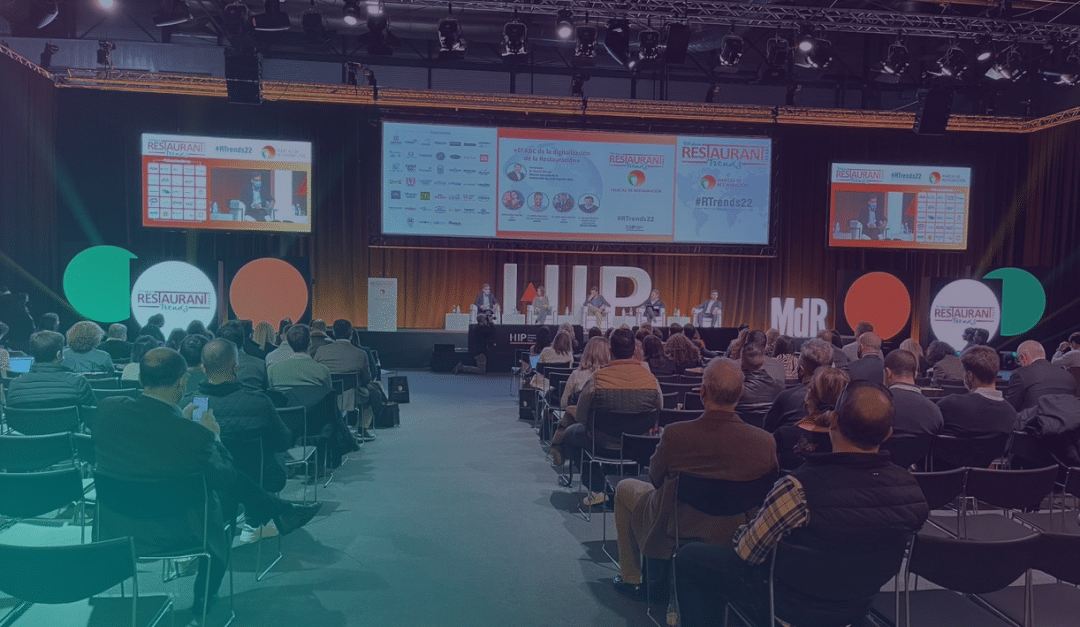 A large audience sits facing a stage where panellists speak at a conference, with presentation slides and banners reading RESTAURANT and event hashtags displayed on screens behind them.