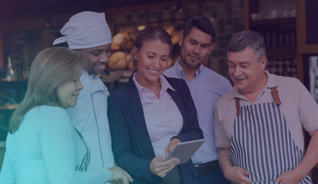 Five adults, including a chef and a person in an apron, stand together in a bakery or café, smiling and looking at a tablet held by a woman in business attire.