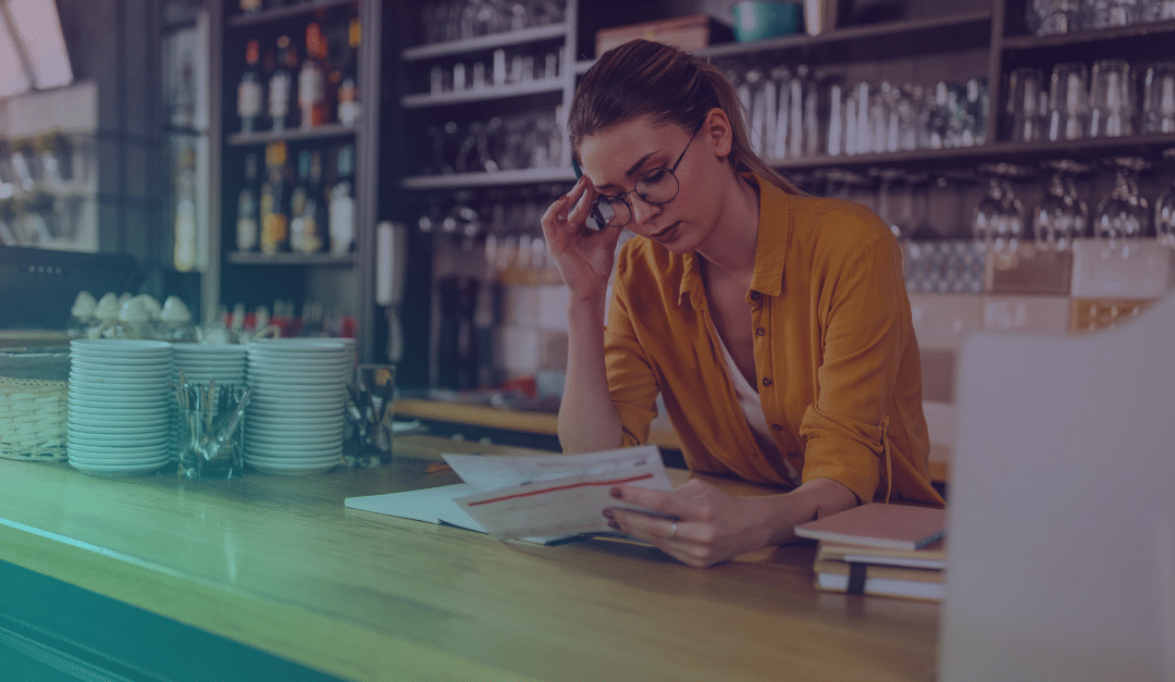 A woman wearing glasses and a yellow shirt reviews paperwork behind a counter in a café or bar, surrounded by cups, glasses, and bottles.