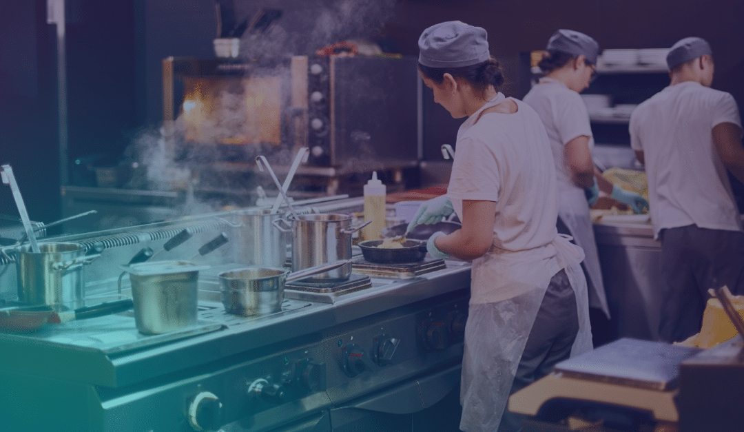 Three chefs in a commercial kitchen work at hobs and prep counters, surrounded by pots, pans, and steam, preparing food in a busy restaurant environment.