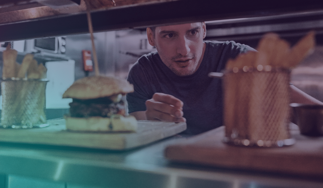A man in a kitchen peers through a serving hatch at a burger and chips served on wooden boards, ready for collection.