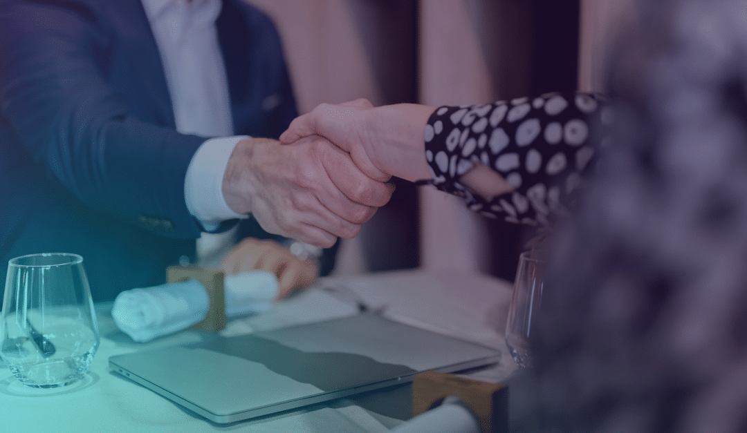 Two people shake hands across a table set with glasses, serviettes, and a closed laptop, suggesting a business meeting or agreement in a formal setting.