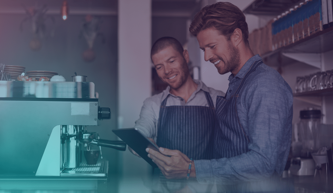 Two men wearing aprons stand beside a coffee machine, smiling as they look at a tablet together in a cosy café setting.