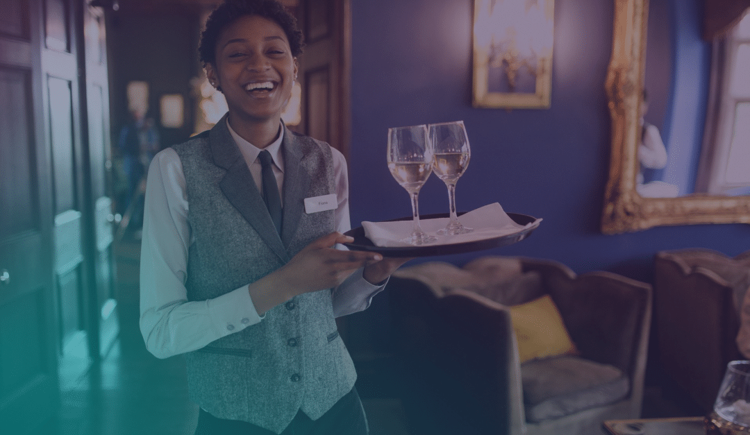 A smiling waiter in a waistcoat and tie holds a tray with two wine glasses in a stylish, warmly lit lounge with framed art and comfortable seating in the background.