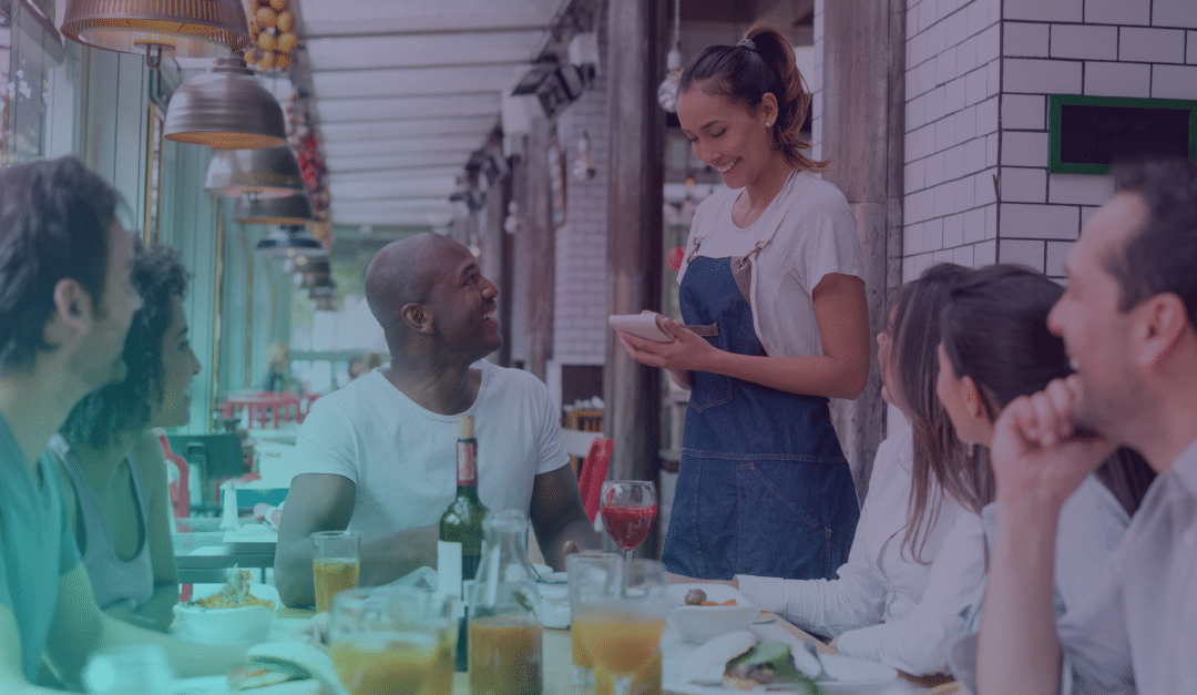 A group of people sit at a restaurant table chatting while a smiling waitress stands, taking their order. Glasses of juice, a wine bottle, and food are on the table. The setting is bright and casual.