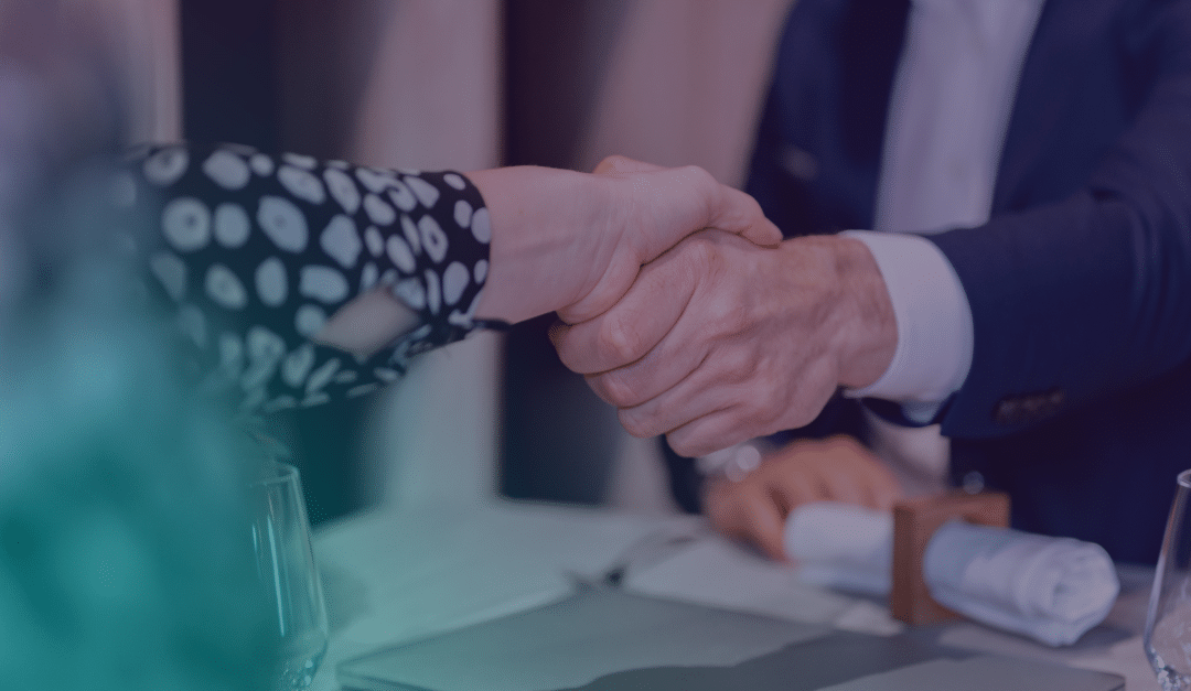 Two people in business attire shake hands across a table, suggesting agreement or partnership, with a glass and a rolled serviette visible in the foreground.