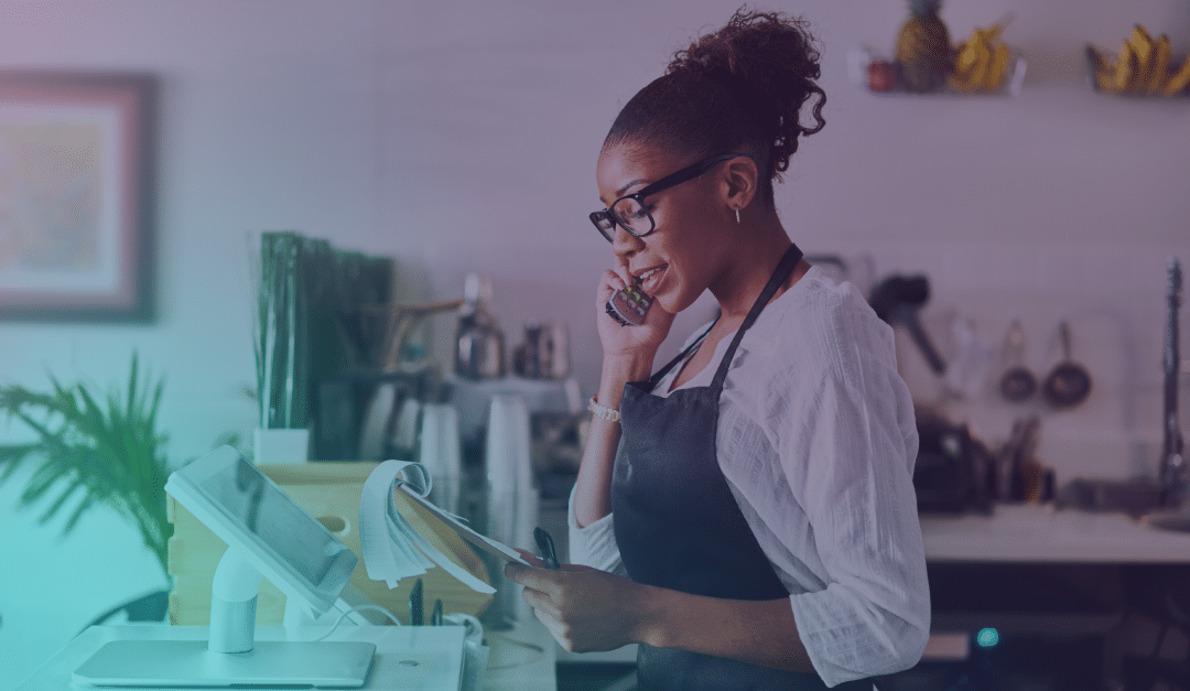 A woman wearing glasses and an apron stands at a counter with a tablet and papers, talking on the phone in a bright, modern kitchen or café.