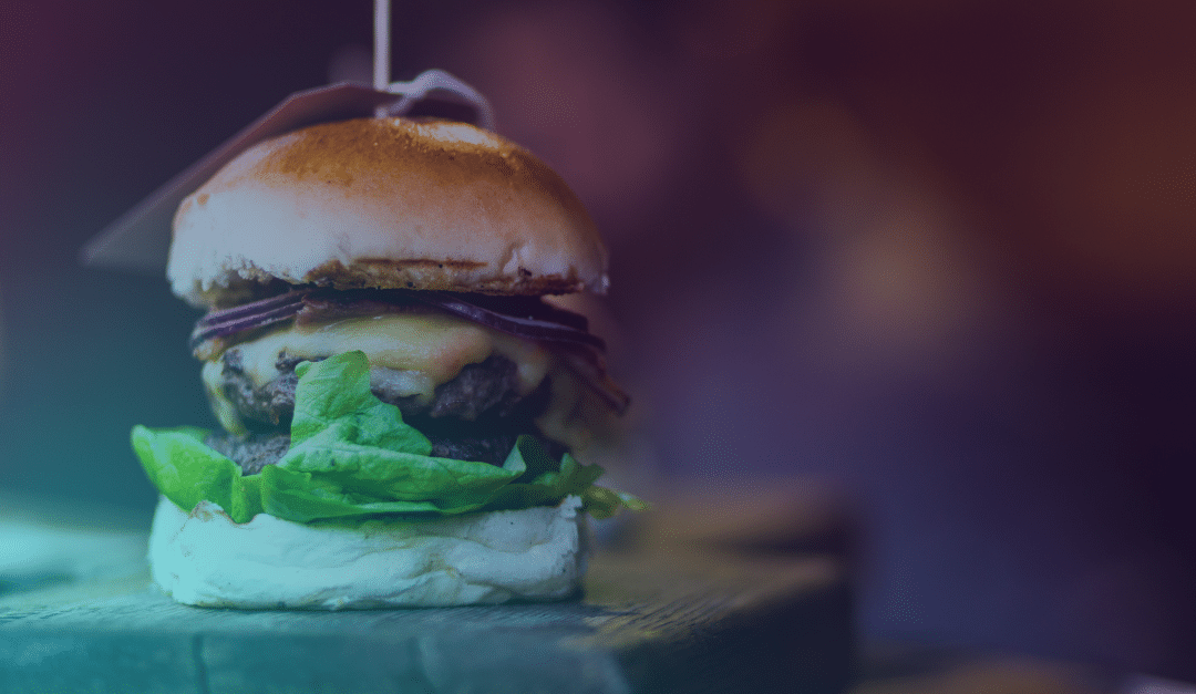 A close-up of a cheeseburger with lettuce, cheese, onions, and a cocktail stick holding it together, set on a wooden surface with a blurred background.