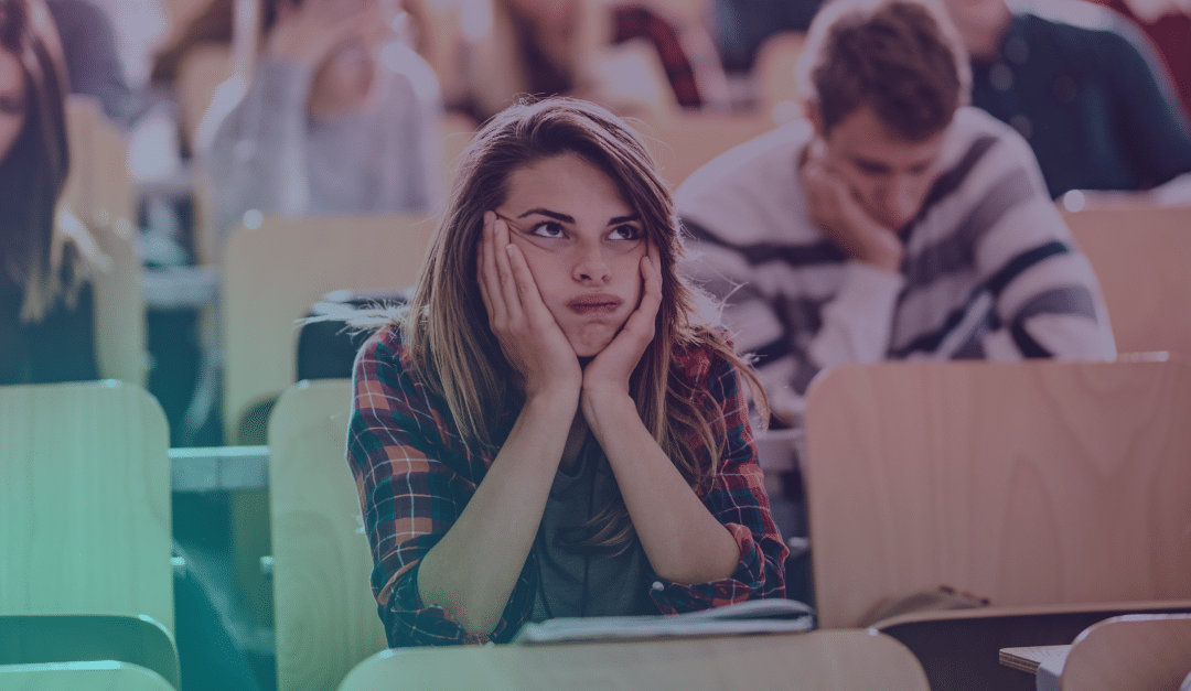 A student in a checked shirt rests her face in her hands, looking bored or frustrated in a lecture theatre, surrounded by other students.