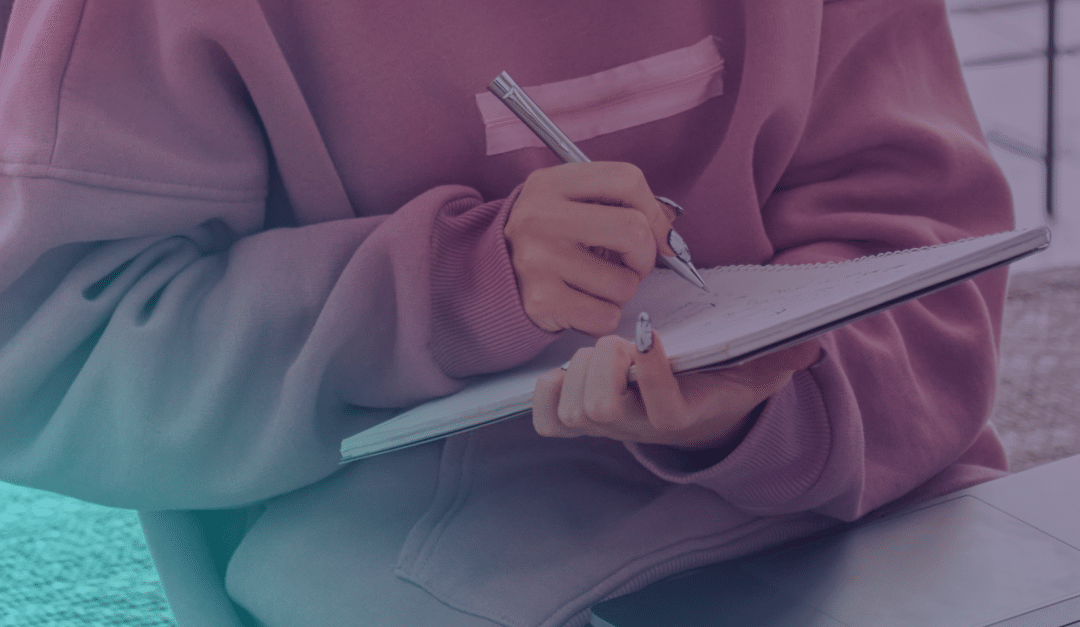 Person wearing a pink jumper writes in a notebook with a pen, sitting next to a laptop on a table. Only their hands and torso are visible.