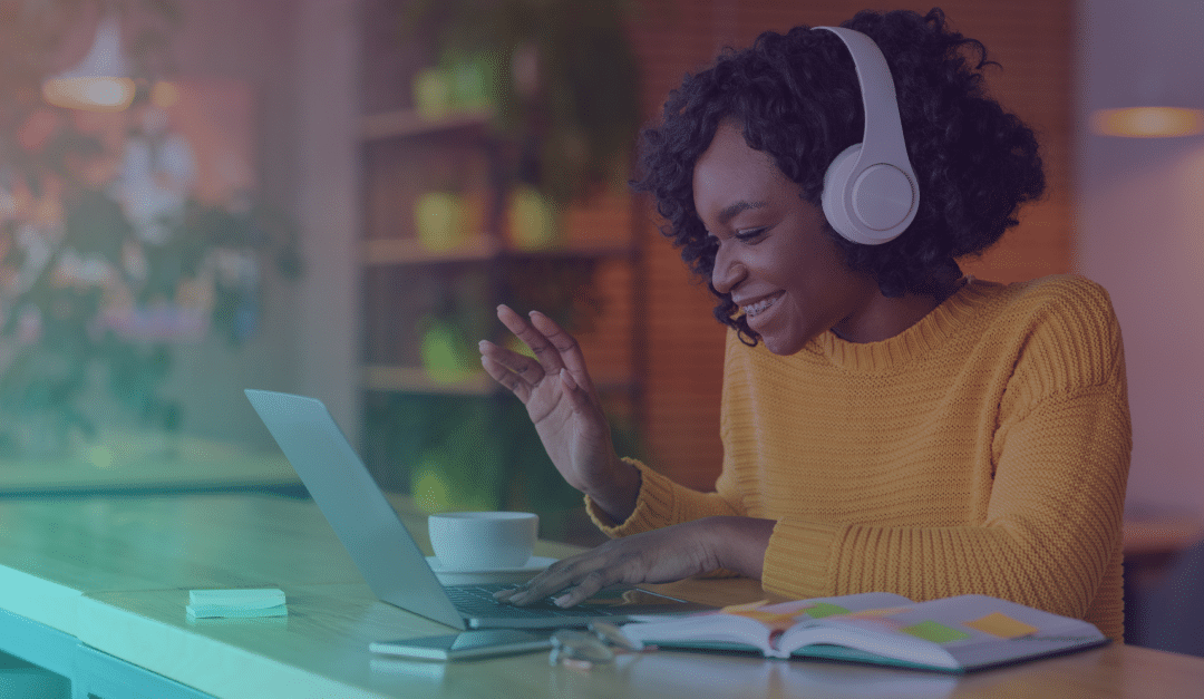 Woman wearing headphones smiles and waves at her laptop during a video call. She sits at a desk with a notebook, pen, and cup, in a cosy, well-lit indoor setting with plants in the background.