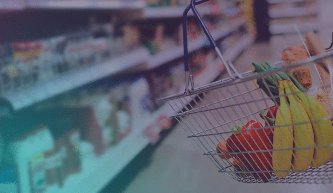 A shopping basket with bananas, bread, and vegetables is held in a supermarket aisle, with shelves of products blurred in the background.
