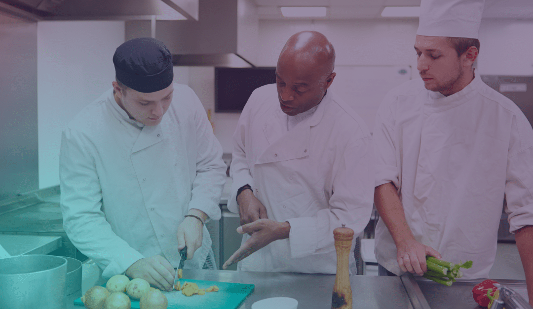 Three chefs in a commercial kitchen; one slices vegetables while another chef gives instructions. The third chef, holding celery, observes attentively.