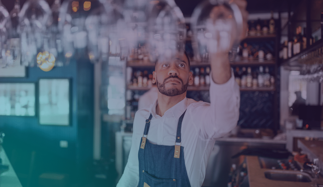 A barman wearing an apron reaches up to grab a wine glass from an overhead rack in a bar filled with bottles and shelves in the background.