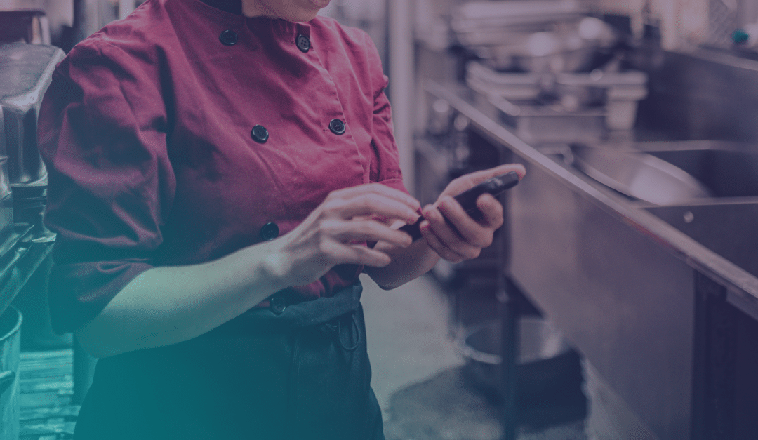 A person in a maroon chef's coat uses a smartphone in a commercial kitchen, standing near metal worktops and industrial sinks.