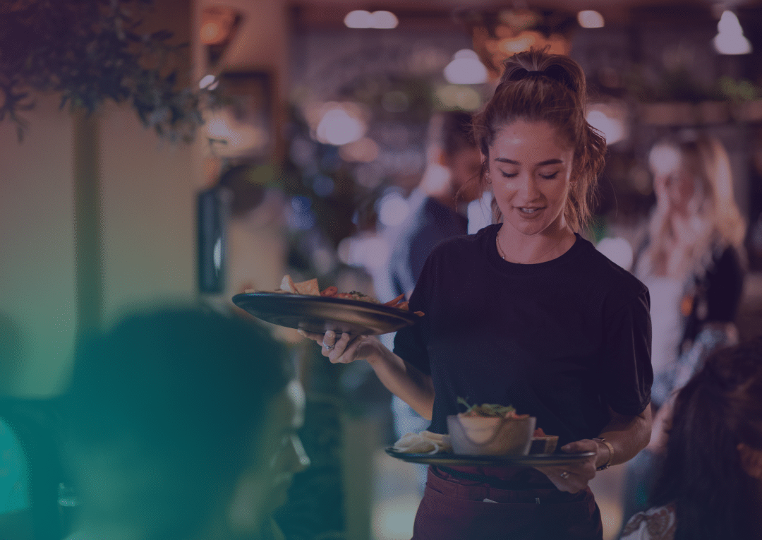 A waiter carrying plates of food to a table in a busy, warmly lit restaurant, with customers blurred in the background.