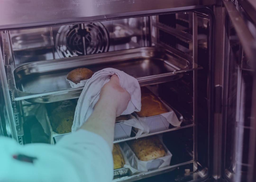 A person wearing a white sleeve removes a baking tray with a muffin from a metal oven, with other loaves in rectangular tins visible on lower shelves.