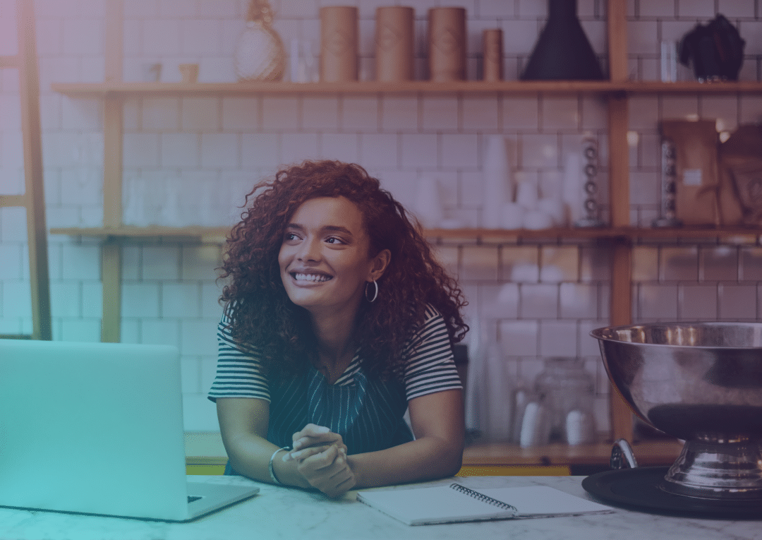 A woman with curly hair and a striped shirt smiles while leaning on a counter in a modern café, with a laptop, notebook, and shelves of jars and cups in the background.