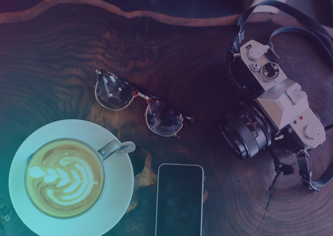 A cup of latte with leaf art, a pair of glasses, a film camera, and a mobile phone rest on a wooden table.