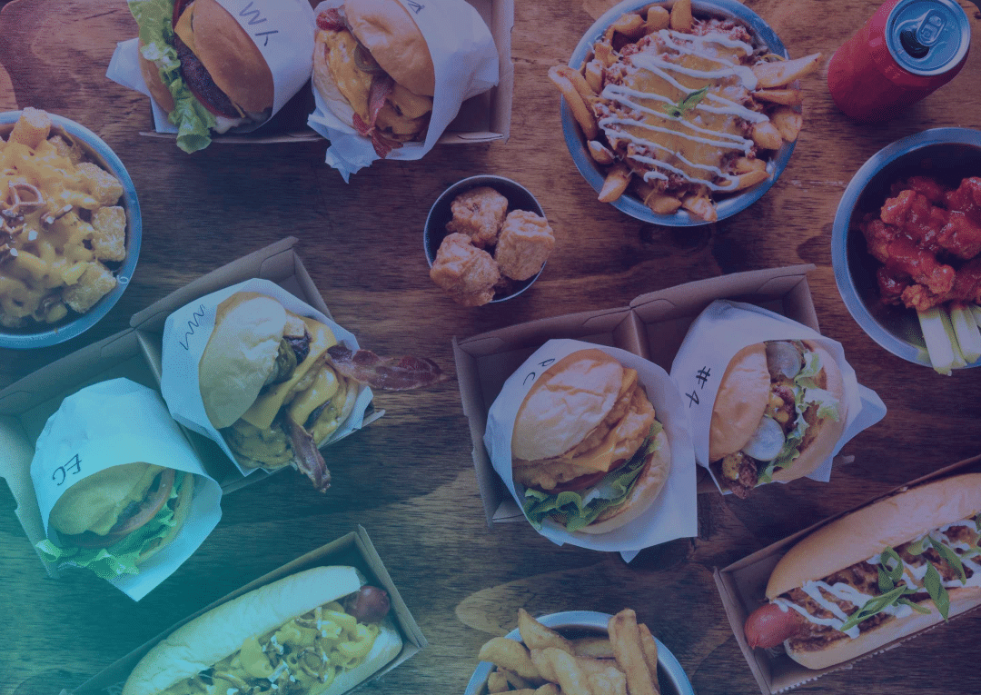 A wooden table covered with burgers, hot dogs, chips, fried chicken wings, macaroni cheese, cheesy chips, a can of fizzy drink, and chicken nuggets in baskets and bowls.