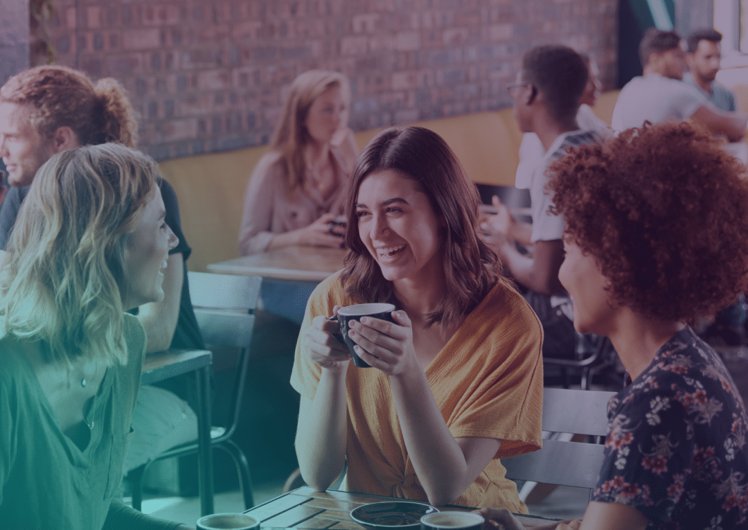 Three women sit at a café table, smiling and chatting over coffee. Other people are talking at tables in the background, creating a lively, friendly atmosphere.