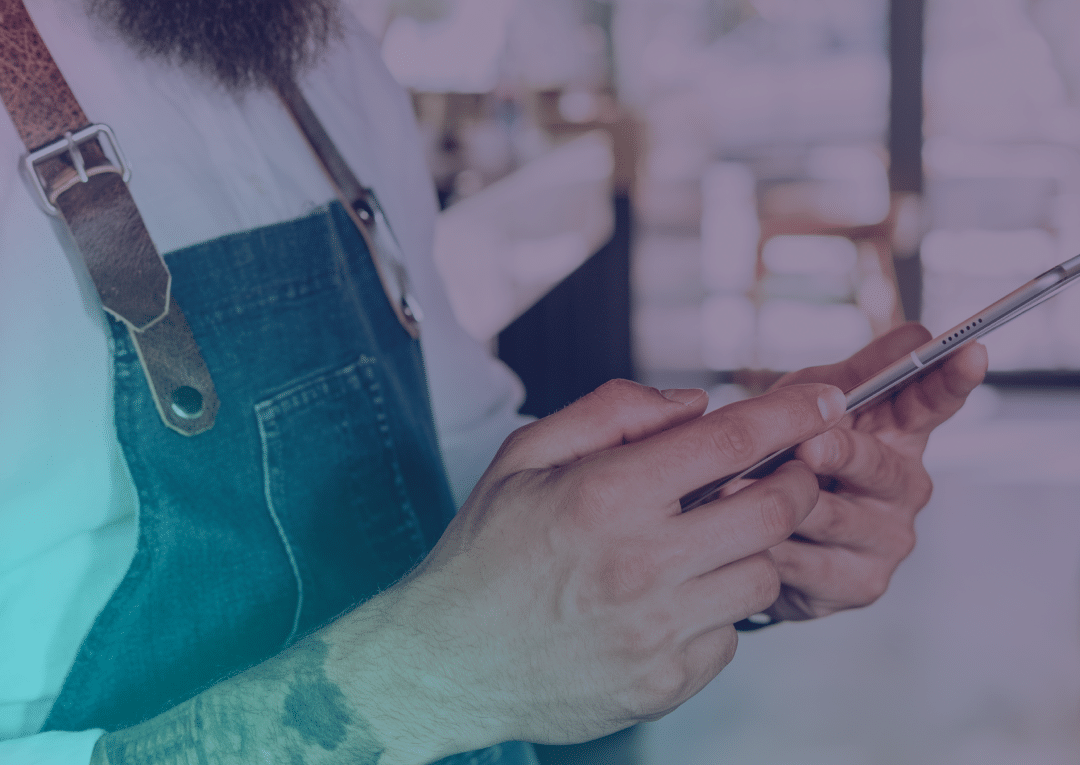 A person wearing a denim apron and white shirt uses a mobile phone, with only their hands, tattoo, and part of their beard visible. The background is out of focus.