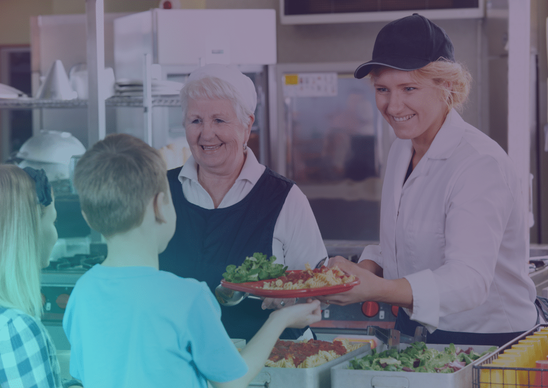 Two canteen workers serve food to two children standing in a lunch queue. One worker hands a plate to a boy while both women smile. The kitchen area and trays of food are visible in the background.