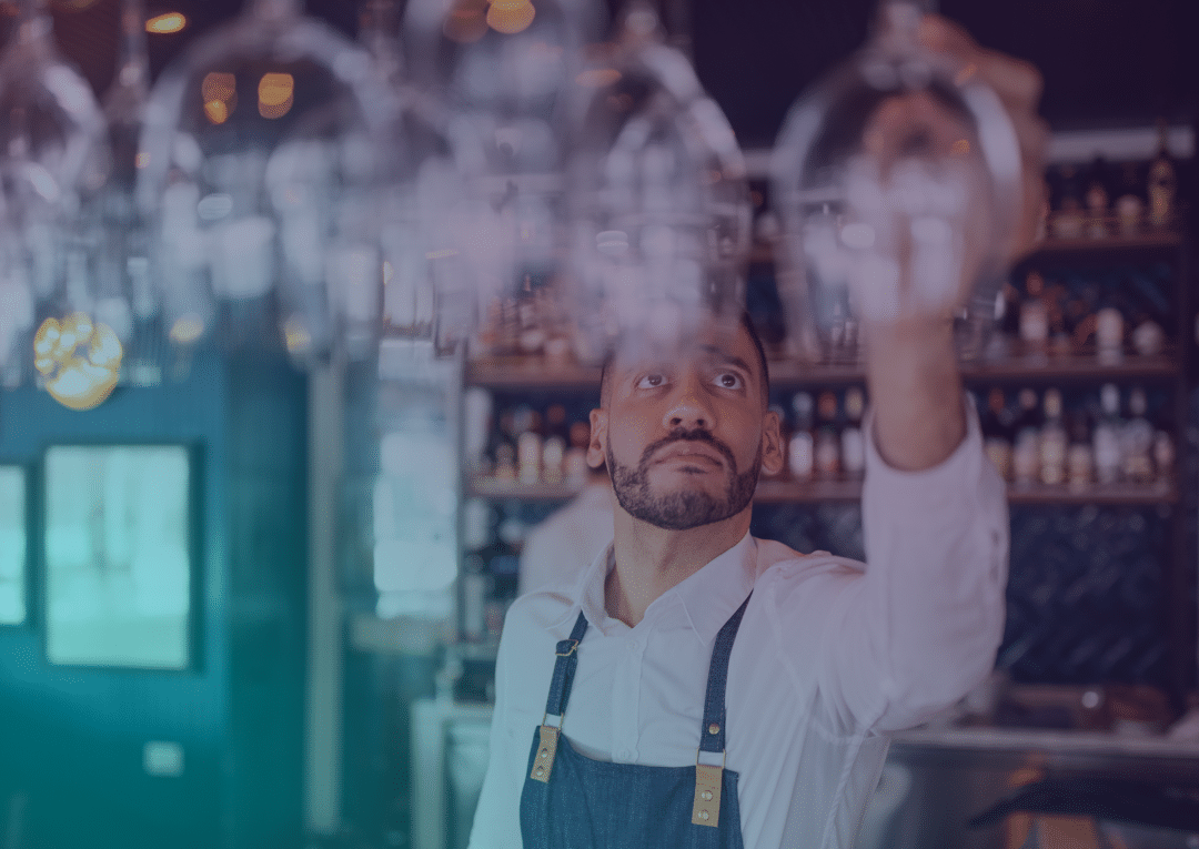 A barman in a white shirt and apron arranges hanging wine glasses in a bar, with shelves of bottles in the background.