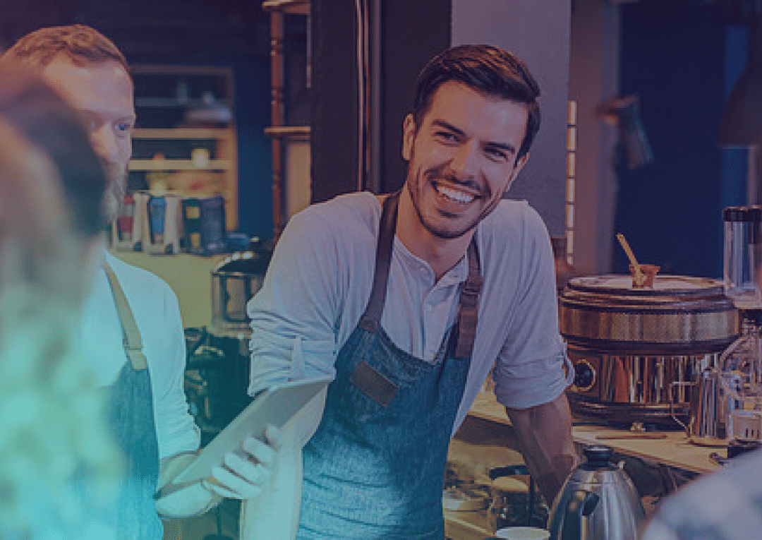 A smiling barista wearing an apron talks with colleagues behind a coffee shop counter, surrounded by equipment like a kettle and mugs.