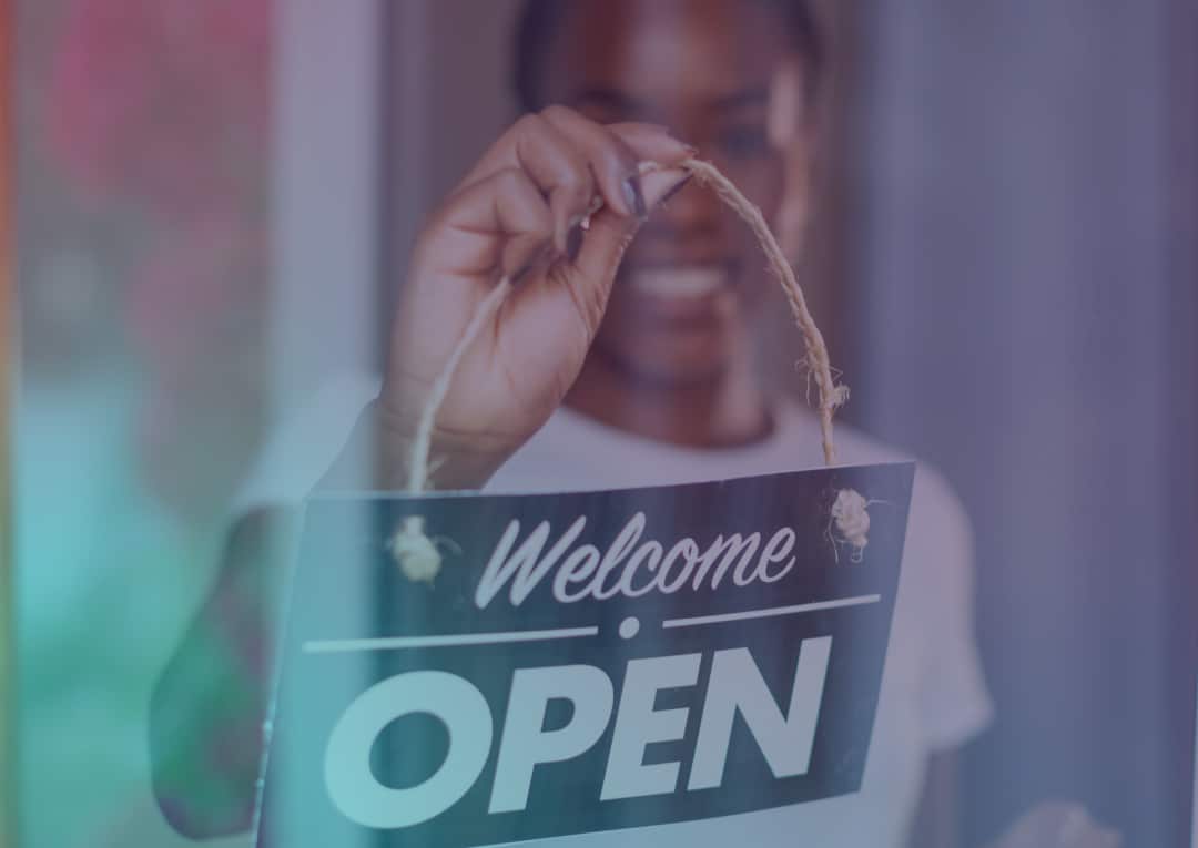 A person smiles while flipping a sign that reads Welcome OPEN on a glass door, indicating the business is now open.