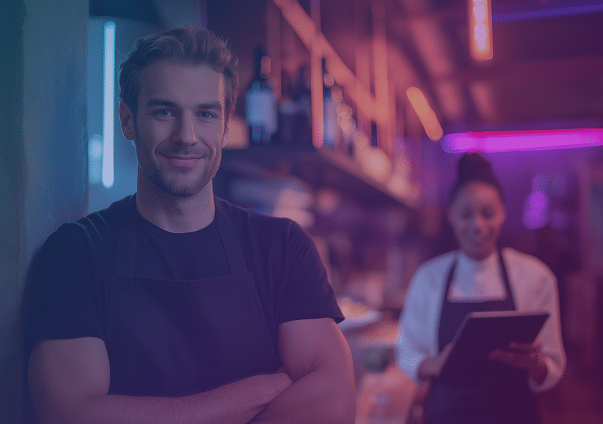 A man in a dark T-shirt and apron stands smiling with arms crossed in a neon-lit restaurant kitchen, while a woman in a white chef's coat holds a tablet in the blurred background.