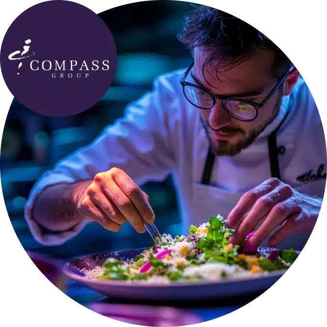 A chef with glasses carefully arranges food on a plate, with the Compass Group logo in the corner. The setting is vibrant and focused on food presentation.