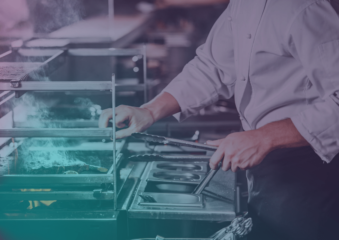 A chef in a white coat uses tongs to cook food on a grill in a commercial kitchen, with steam rising from the cooking surface.
