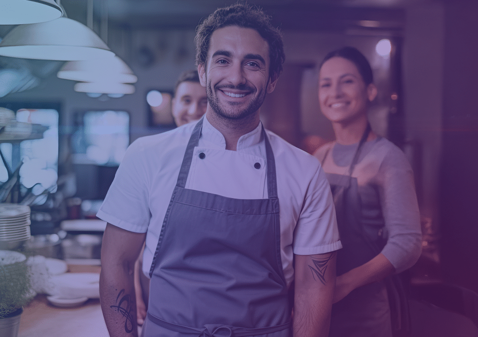 A smiling man in a chef's uniform stands in a restaurant kitchen, with two colleagues in aprons in the background. They look happy and welcoming.
