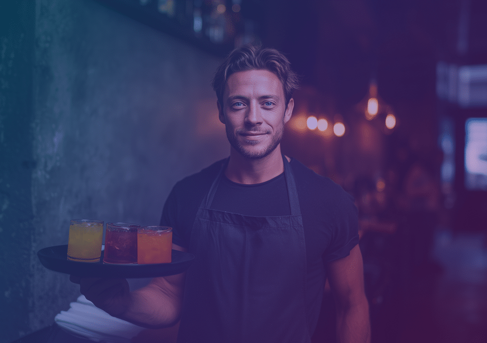 A smiling barman wearing a black apron holds a tray with colourful drinks in a dimly lit bar, with warm lights blurred in the background.