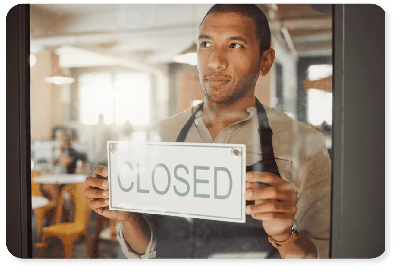 A person wearing an apron stands inside a café, holding a CLOSED sign against the glass door, looking thoughtfully outside.