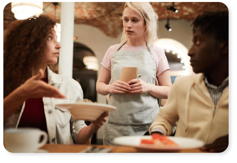 A woman holding a plate gestures towards a waitress standing by the table, while another person with food sits beside her. The scene appears to be a restaurant with a serious or confused atmosphere.