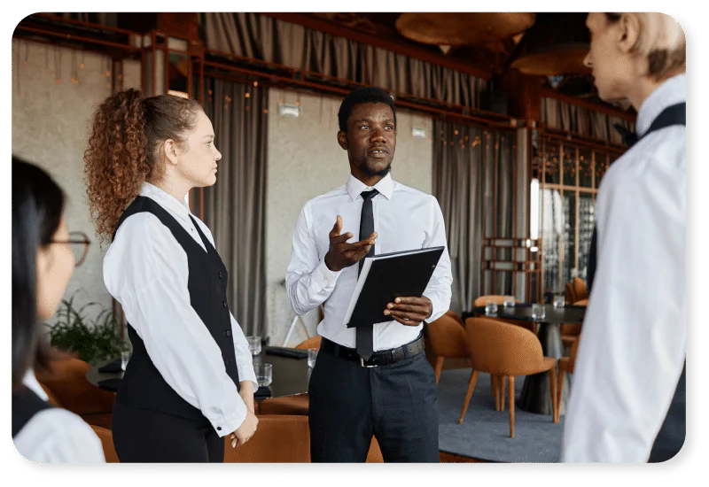 A man holding a clipboard speaks to three colleagues in uniforms, likely restaurant staff, during a meeting in a stylish dining area with brown chairs and large windows.